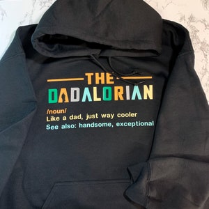 Op de afbeelding: Zwart hoodie met een kleurrijk grafisch ontwerp met de tekst "The Dadalorian /noun/ Like a dad, just way cooler See also: handsome, exceptional"