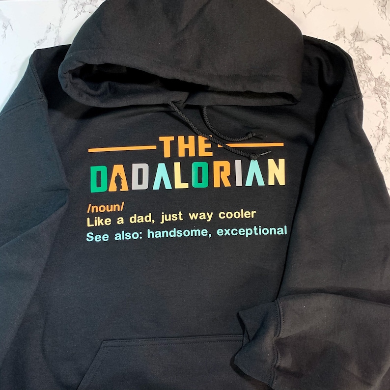 Op de afbeelding: Zwart hoodie met een kleurrijk grafisch ontwerp met de tekst "The Dadalorian /noun/ Like a dad, just way cooler See also: handsome, exceptional"