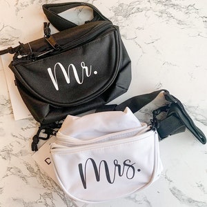 Op de afbeelding: Twee heuptasjes, &eacute;&eacute;n zwart en &eacute;&eacute;n wit, worden getoond. De zwarte tas heeft "Mr." in witte letters en de witte tas heeft "Mrs." in zwarte letters. Beide hebben verstelbare bandjes en ritsen.