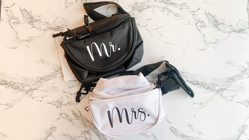 Op de afbeelding: Twee heuptasjes, &eacute;&eacute;n zwart en &eacute;&eacute;n wit, worden getoond. De zwarte tas heeft "Mr." in witte letters en de witte tas heeft "Mrs." in zwarte letters. Beide hebben verstelbare bandjes en ritsen.