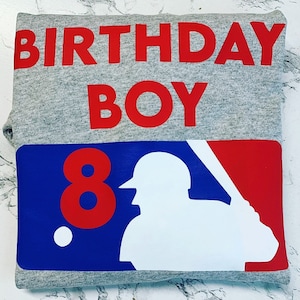 May include: A gray sweatshirt with the words "BIRTHDAY BOY" in red. Below, a baseball-themed graphic with the number "8" in red, a white baseball player silhouette, and a white baseball on a blue and red background.