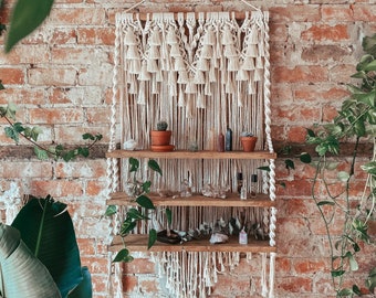May include: A macrame wall hanging with a wooden shelf and three tiers. The shelf is decorated with small potted plants, crystals, and bottles. The wall hanging is cream coloured and has a fringe.