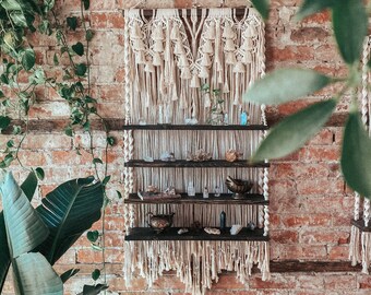 May include: A cream macrame wall shelf with three dark wooden shelves, displaying crystals and small ornaments. The shelf hangs against a red brick wall. Green plants are visible on either side.