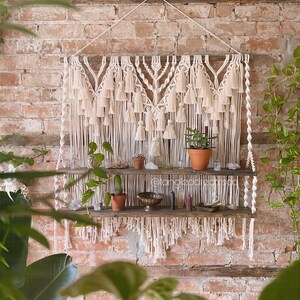 Op de afbeelding: Een macramé wandhanger met een geometrisch ontwerp en kwastjes hangt boven een houten plank met twee niveaus. De plank is versierd met potplanten, kristallen en andere decoratieve items. De muur is van baksteen.