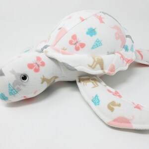 May include: A plush sea turtle toy made of white fabric featuring a repeating pattern of pink butterflies, blue trees, and tan kangaroos. The stuffed animal has a rounded shell, four flippers, and a head with a single visible black plastic eye.