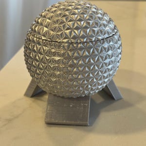 May include: A silver-colored, spherical decorative box with a geometric pattern. The box is supported by a three-legged stand. The box has a lid that appears to be removable. The box is made of a metallic material.