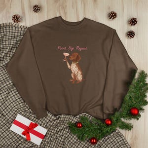 May include: A brown sweatshirt featuring a dog illustration holding a wine glass, with the text "Point. Sip. Repeat." The dog has brown and white markings and a green collar. The image is decorated with pine cones and red baubles.