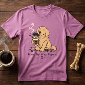 May include: A lilac t-shirt with a cartoon golden retriever dog holding a coffee cup. The dog is smiling. The text "Brew. Sip. Wag. Repeat." is printed below the dog. A coffee cup and a dog bone are on the sides, all on a wooden surface.