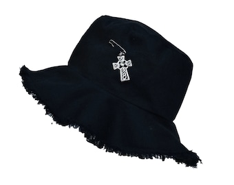 Könnte beinhalten: Schwarzer Bucket Hat mit ausgefransten Kanten und einem silbernen keltischen Kreuz-Anhänger, der mit einer Sicherheitsnadel befestigt ist.