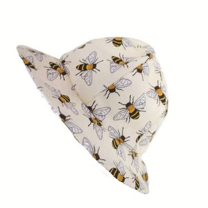 May include: A white bucket hat with a pattern of yellow and black bees. The hat has a wide brim and a rounded crown.