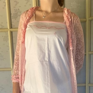 Puede incluir: Una bata de encaje rosa con un camisón de algodón blanco y pantalones cortos debajo. El camisón y los pantalones cortos tienen ribetes y bordados rosas.