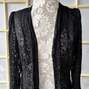 May include: A black lace duster coat with long sleeves and a flowing design. The coat is displayed on a mannequin with a white lace top, showcasing the intricate floral pattern of the lace fabric. The coat has a classic, elegant style.