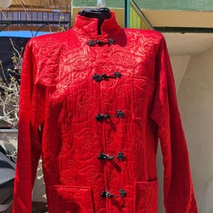 May include: A red quilted jacket with a mandarin collar and black button closures. The jacket has a floral pattern and two pockets.