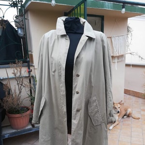 Op de afbeelding: Een beige lange jas met lange mouwen, een knoopsluiting en één zak aan de linkerkant. De jas is op een mannequin en wordt getoond tegen een betegelde achtergrond.