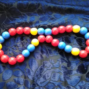May include: A colorful beaded necklace with red, blue, and yellow beads. The necklace is arranged in a circle shape and is lying on a blue fabric background.