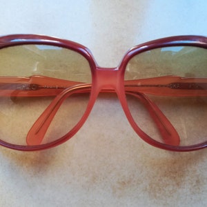 Könnte beinhalten: Eine Sonnenbrille mit rotem Kunststoffgestell und braunen Gläsern. Die Sonnenbrille hat den Text "O.H.A. 5118" auf dem rechten Bügel.