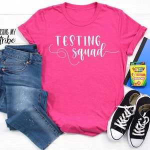 Könnte beinhalten: Pinkes T-Shirt mit dem Text "Testing Squad" in weißen Buchstaben. Das Shirt wird mit einer blauen Jeans, schwarzen Sneakern und einer Schachtel mit Buntstiften kombiniert.