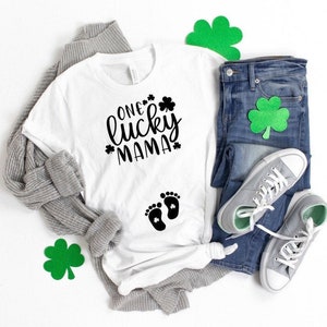 May include: White t-shirt with black text that reads "One lucky mama" and a pair of black baby footprints with a shamrock inside each footprint. The shirt is laid out on a white surface with a gray cardigan, blue jeans, and green shamrock decorations.
