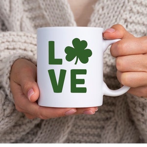 Op de afbeelding: Een witte keramische mok met een groene klaver en het woord "LOVE" in groene letters.