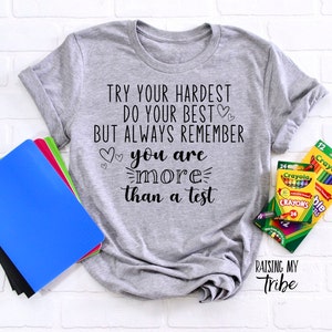 May include: Gray t-shirt with the text "Try your hardest do your best but always remember you are more than a test" and two hearts. The shirt is laying on a white brick background with school supplies.