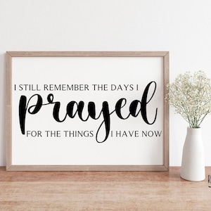 May include: A framed print with a black and white inspirational quote that reads "I still remember the days I prayed for the things I have now." The quote is in a cursive font. The frame is light brown wood. The print is sitting on a wooden surface. Below the print is a white vase with white flowers. The text "Raising My Tribe" is printed in black below the vase.