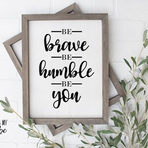 May include: A framed sign with the text "Be brave, be humble, be you" in black script font on a white background. The sign is in a gray wooden frame and is sitting on a white brick wall.