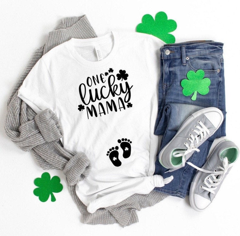 May include: White t-shirt with black text that reads "One lucky mama" and a pair of black baby footprints with a shamrock inside each footprint. The shirt is laid out on a white surface with a gray cardigan, blue jeans, and green shamrock decorations.