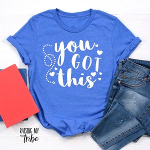 May include: A royal blue t-shirt with the text "you got this" in white with small white hearts around the text. The t-shirt is laying on a white surface with a pair of blue jeans and a red book.
