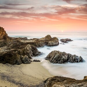 Könnte beinhalten: Ein felsiger Strand mit einem sandigen Ufer bei Sonnenuntergang. Der Himmel ist in leuchtendem Rosa und Orange gefärbt, und die Wellen schlagen gegen die Felsen.