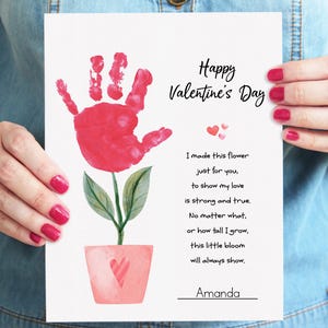 May include: A Valentine's Day card featuring a red handprint flower with green leaves and a pink pot. The card includes the text "Happy Valentine's Day" and a poem. The card is held by a person with red nail polish.