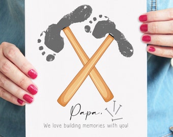 Op de afbeelding: Een witte kaart met een grijs voetafdruk-ontwerp dat eruitziet als een hamer. De kaart zegt "Papa. We houden ervan om herinneringen met je te maken!"