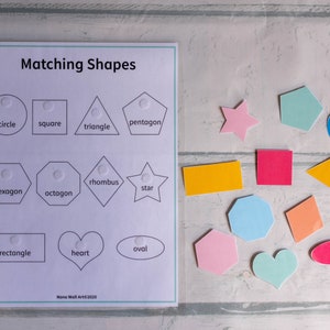 Op de afbeelding: Een wit vel papier met de titel "Matching Shapes" bovenaan gedrukt.  Het vel is verdeeld in drie rijen met vier vormen in elke rij.  Elke vorm heeft een wit puntje in het midden.  De vormen zijn gelabeld met de volgende woorden: cirkel, vierkant, driehoek, vijfhoek, zeshoek, achthoek, ruit, ster, rechthoek, hart en ovaal.  Onder de vormen staat de tekst "Nono Wall Art&copy;2020".  Er zijn 12 kleurrijke vormen op een wit houten oppervlak.  De vormen zijn: een ster, een cirkel, een vierkant, een vijfhoek, een zeshoek, een achthoek, een ruit, een rechthoek, een hart, een ovaal, een driehoek en een ruit.