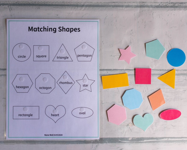 Op de afbeelding: Een wit vel papier met de titel "Matching Shapes" bovenaan gedrukt.  Het vel is verdeeld in drie rijen met vier vormen in elke rij.  Elke vorm heeft een wit puntje in het midden.  De vormen zijn gelabeld met de volgende woorden: cirkel, vierkant, driehoek, vijfhoek, zeshoek, achthoek, ruit, ster, rechthoek, hart en ovaal.  Onder de vormen staat de tekst "Nono Wall Art&copy;2020".  Er zijn 12 kleurrijke vormen op een wit houten oppervlak.  De vormen zijn: een ster, een cirkel, een vierkant, een vijfhoek, een zeshoek, een achthoek, een ruit, een rechthoek, een hart, een ovaal, een driehoek en een ruit.
