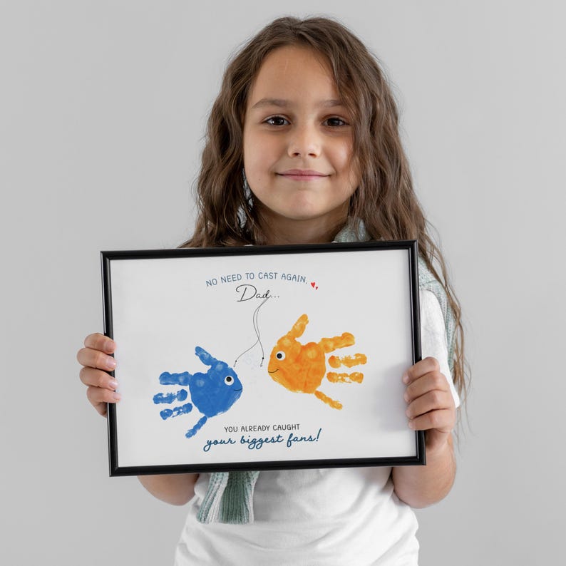 May include: A framed print with handprint fish art, one blue and one orange, linked by a line. Text says "NO NEED TO CAST AGAIN, Dad..." and "YOU ALREADY CAUGHT your biggest fans!" The frame is black.