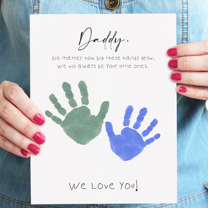 Printable Father's Day handprint art held by a mom showing two children's handprints in sage green and blue with the message "Daddy, no matter how big these hands grow, we will always be your little ones — we love you" in mixed script and print lettering on white