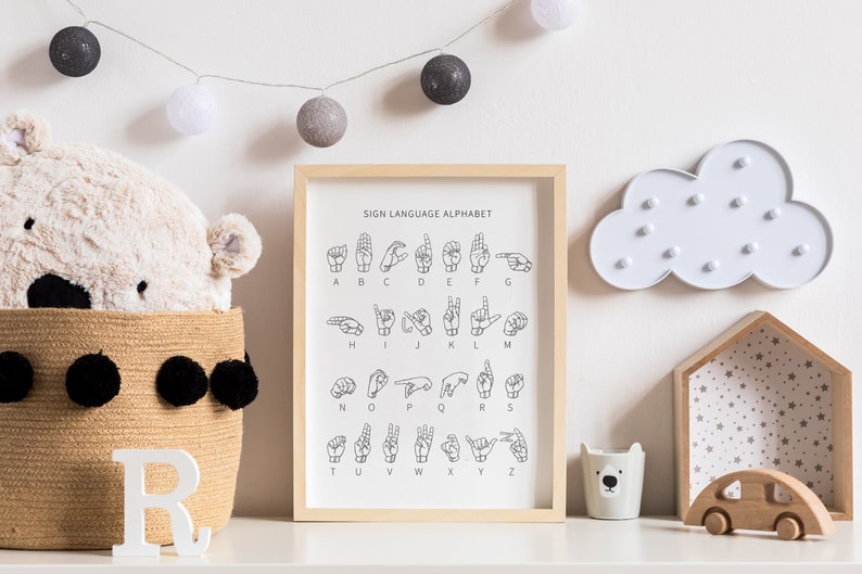 May include: A framed print of the American Sign Language alphabet with a white background and black lettering. The print is hanging on a white wall with a string of white and gray cotton ball lights above it. There is a stuffed animal, a basket, and a wooden toy car on a white shelf below the print.