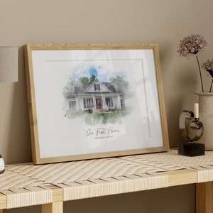 Puede incluir: Una impresi&oacute;n en acuarela enmarcada de una casa blanca con un marco de madera marr&oacute;n claro. La impresi&oacute;n est&aacute; etiquetada como "Our First Home" con la direcci&oacute;n debajo. Una l&aacute;mpara, un jarr&oacute;n con flores y un objeto decorativo est&aacute;n sobre una mesa tejida.