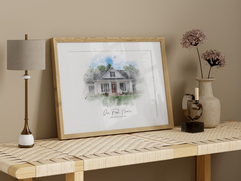 Puede incluir: Una impresi&oacute;n en acuarela enmarcada de una casa blanca con un marco de madera marr&oacute;n claro. La impresi&oacute;n est&aacute; etiquetada como "Our First Home" con la direcci&oacute;n debajo. Una l&aacute;mpara, un jarr&oacute;n con flores y un objeto decorativo est&aacute;n sobre una mesa tejida.