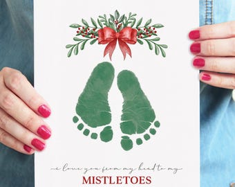 May include: A white rectangular card featuring a watercolor illustration of green baby footprints centered below a red bow and green mistletoe sprigs. The text reads, I love you from my head to my MISTLETOES, with MISTLETOES in red ink.
