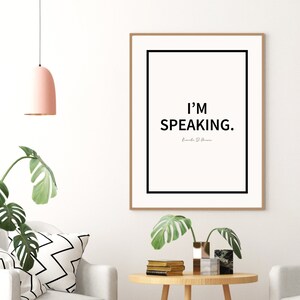 May include: A black and white framed print with the text "I'M SPEAKING." and the name "Emily D. Herrera" in a smaller font below. The print is hanging on a white wall.