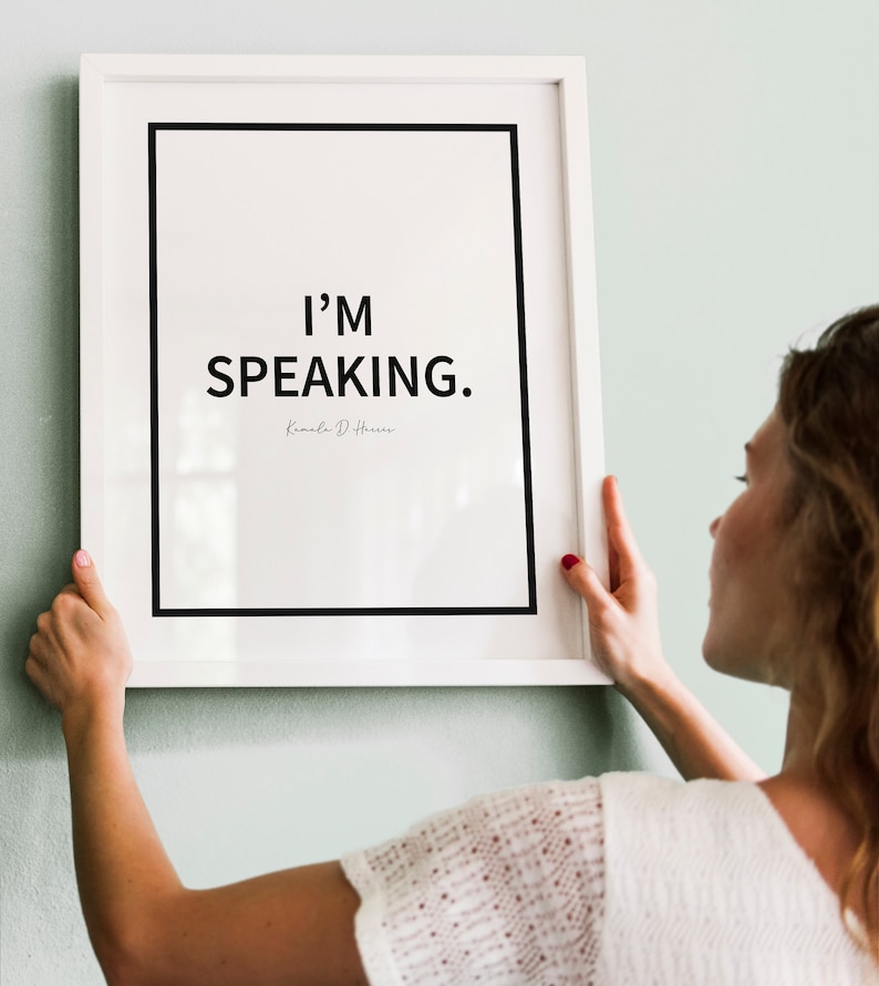 May include: A white framed print with the text "I'M SPEAKING." and the name "Kamala D. Harris" in cursive below. A person is hanging the print on a light green wall.