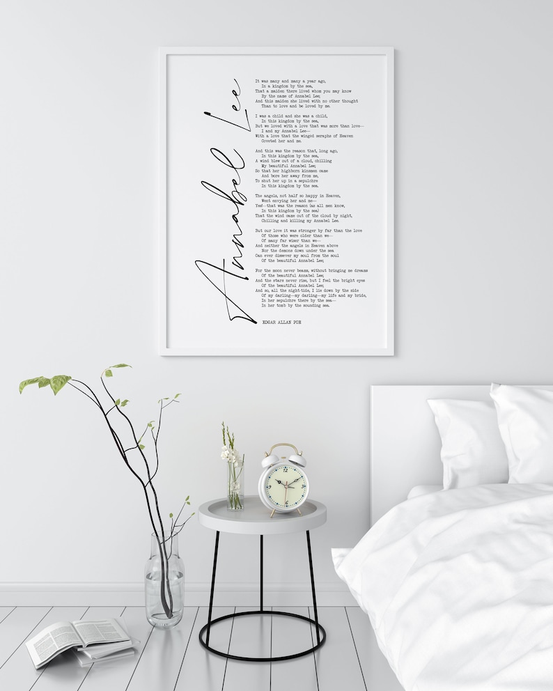 May include: A white framed print of a poem titled "Annabel Lee" by Edgar Allan Poe. The poem is printed in black ink on a white background. The print is hanging on a white wall above a white nightstand with a white alarm clock.
