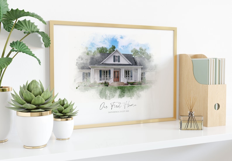 Puede incluir: Una impresi&oacute;n en acuarela enmarcada de una casa blanca con techo negro y puerta marr&oacute;n. La impresi&oacute;n est&aacute; en un marco dorado y tiene el texto "Our First Home" y "Santa Monica, CA EST 2020". La impresi&oacute;n se exhibe en un estante blanco con plantas.