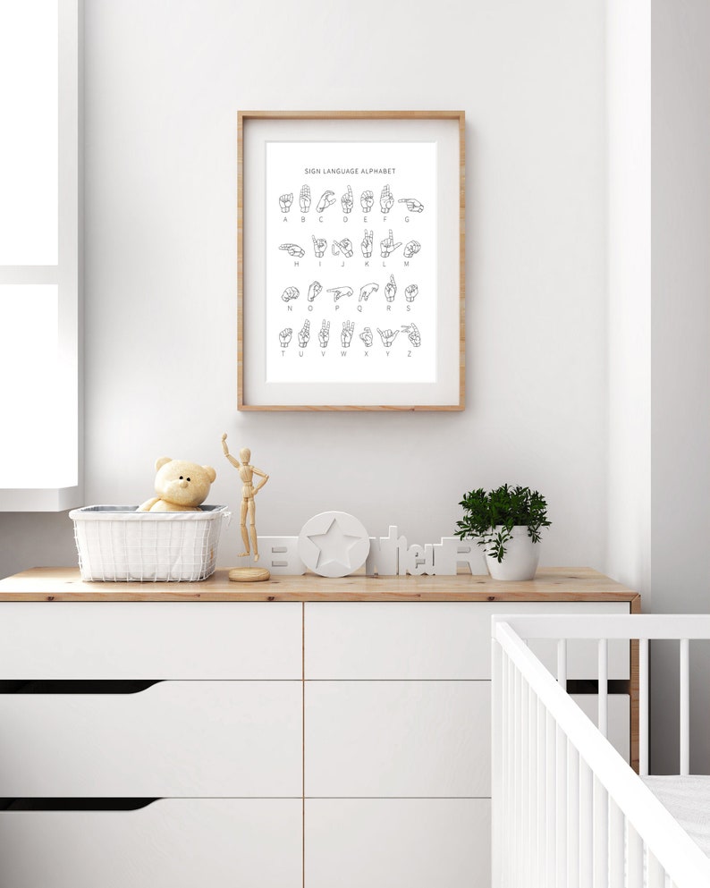 May include: A black and white illustration of the American Sign Language alphabet. The illustration is framed and hanging on a white wall. The wall is in a nursery with a white dresser, a crib, and a teddy bear.