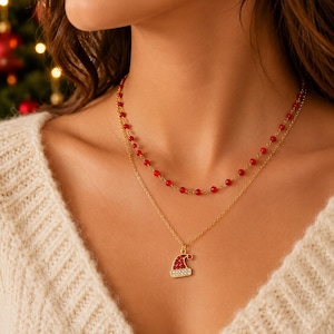 May include: A layered gold-tone necklace featuring two chains. The shorter chain is adorned with small red beads, while the longer chain holds a pendant shaped like a Santa hat, embellished with red and clear crystals set in a gold-tone metal frame.