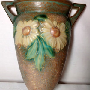 Può includere: Vaso decorativo vintage con finitura strutturata e maculata. Il vaso presenta due manici ed è decorato con margherite in rilievo nei colori crema, giallo e verde. Il vaso ha una combinazione di colori marrone e verde.