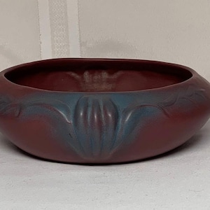 May include: A low, round ceramic bowl with a matte dark red finish. The exterior features a raised, molded relief pattern of stylized leaves and central floral elements highlighted with a subtle blue-grey glaze accenting the curves.