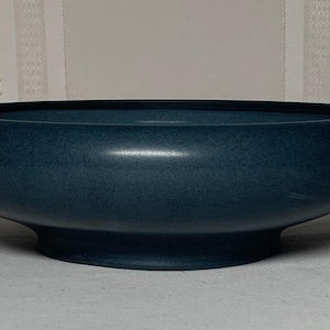 May include: A wide, shallow bowl with a matte, deep teal finish. The bowl features a rounded, curved rim that tapers inward toward a circular base, creating a smooth, continuous silhouette with a consistent dark blue-green color throughout.