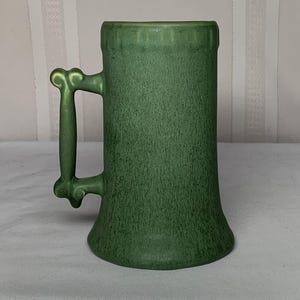 Puede incluir: Una jarra de cerveza cilíndrica de color verde con un acabado mate texturizado y un asa distintiva con forma de hueso. La jarra presenta una base ligeramente ensanchada y un borde liso y redondeado, mostrando un color verde profundo uniforme.