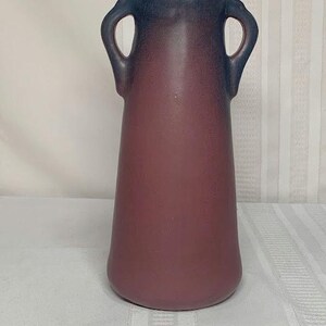 May include: A tall, maroon-colored vase with a flared base and two loop handles near the top. The top of the vase is a darker shade of blue. The vase has a smooth, matte finish and is likely ceramic.